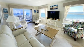 A spacious living room with a sofa, armchairs, a coffee table, and a TV. There are large windows with a view of the sea.