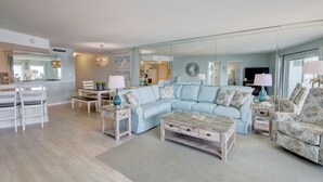 A modern living room with a light blue sectional sofa, a wooden coffee table, and a dining area with white chairs and a small table. - 554 El Matador 1 Bedroom Condo by RedAwning (Fort Walton Beach)