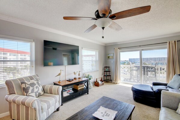 A spacious living room with a ceiling fan, a flat-screen TV, a sofa, a coffee table, and a balcony with a view of buildings. - 440 El Matador 2 Bedroom Condo by RedAwning (Fort Walton Beach)