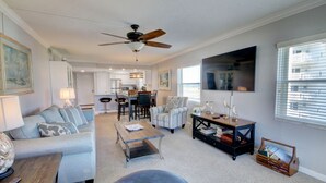 A spacious living room with a sofa, coffee table, and armchair, a dining area with a table and chairs, a kitchen in the background, and a large TV on the wall.