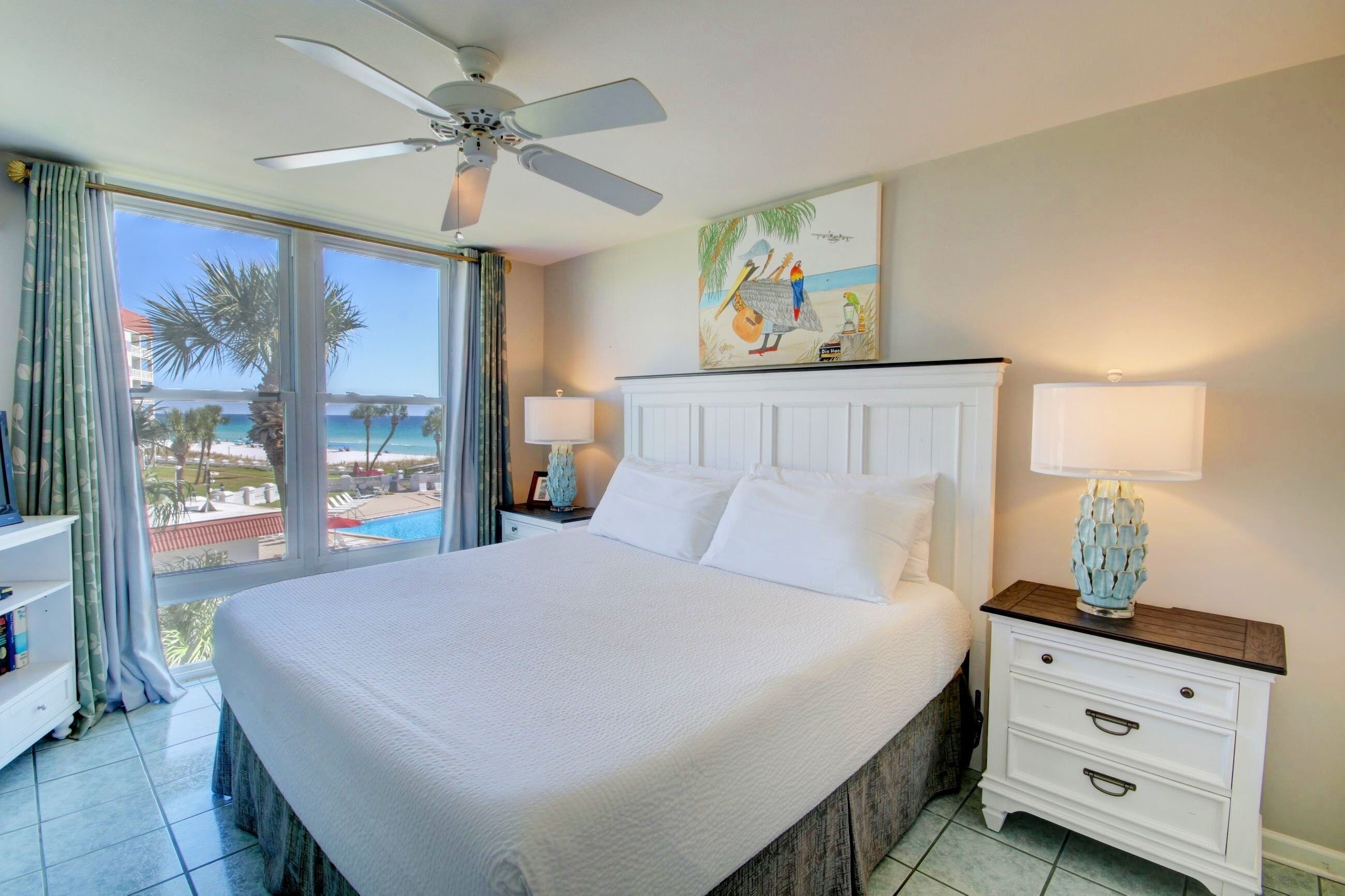 A hotel room with a large bed, a ceiling fan, a nightstand with a lamp, a painting of birds, and a view of the beach and palm trees.