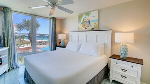Une chambre d’hôtel avec un grand lit, un ventilateur de plafond, une table de chevet avec une lampe, un tableau représentant des oiseaux et une vue sur la plage et les palmiers.