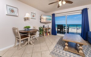 A dining area with a table and chairs, a ceiling fan, a bookshelf, and a large window with a view of the beach. - Tradewinds 203 1 Bedroom Condo (Orange Beach)
