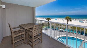A balcony with a wooden table and chairs, a pool, and a beach view.