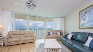 Ein geräumiges Wohnzimmer mit einem beigefarbenen Ecksofa, einem dunkelgrünen Sofa und einem weißen Couchtisch. Ein großes Fenster bietet Blick auf die Skyline der Stadt.