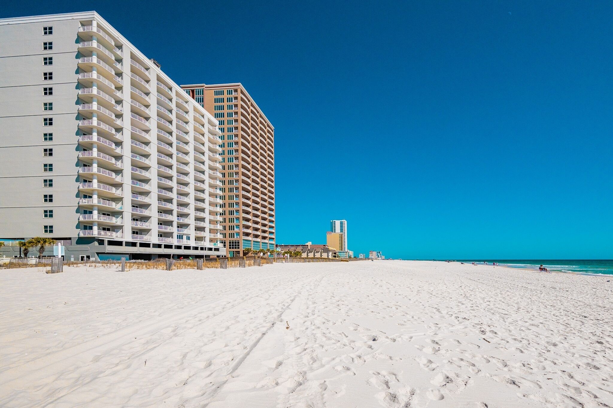 Ein Hotel direkt am Strand mit mehreren Etagen und Balkonen, ein sandiger Strand im Vordergrund und ein klarer, blauer Himmel.