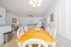 A dining area with a wooden table, white chairs, a ceiling fan, and a window with white shutters.