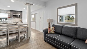 Une cuisine moderne avec des meubles blancs et un îlot central, un canapé gris agrémenté de coussins à motifs et une fenêtre avec des stores.