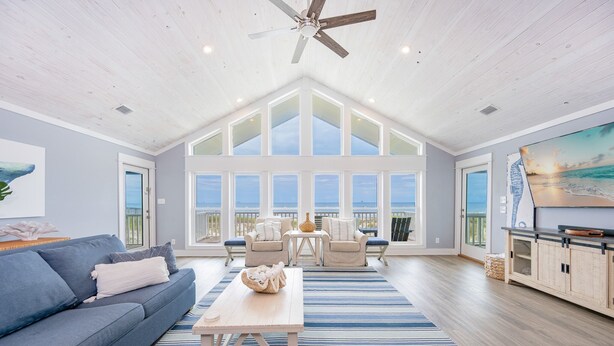 A spacious living room with a high ceiling, large windows, a ceiling fan, a blue sofa, a coffee table, and a view of the ocean. - Blue Hideaway 2888 3 Bedroom Home (Gulf Shores)