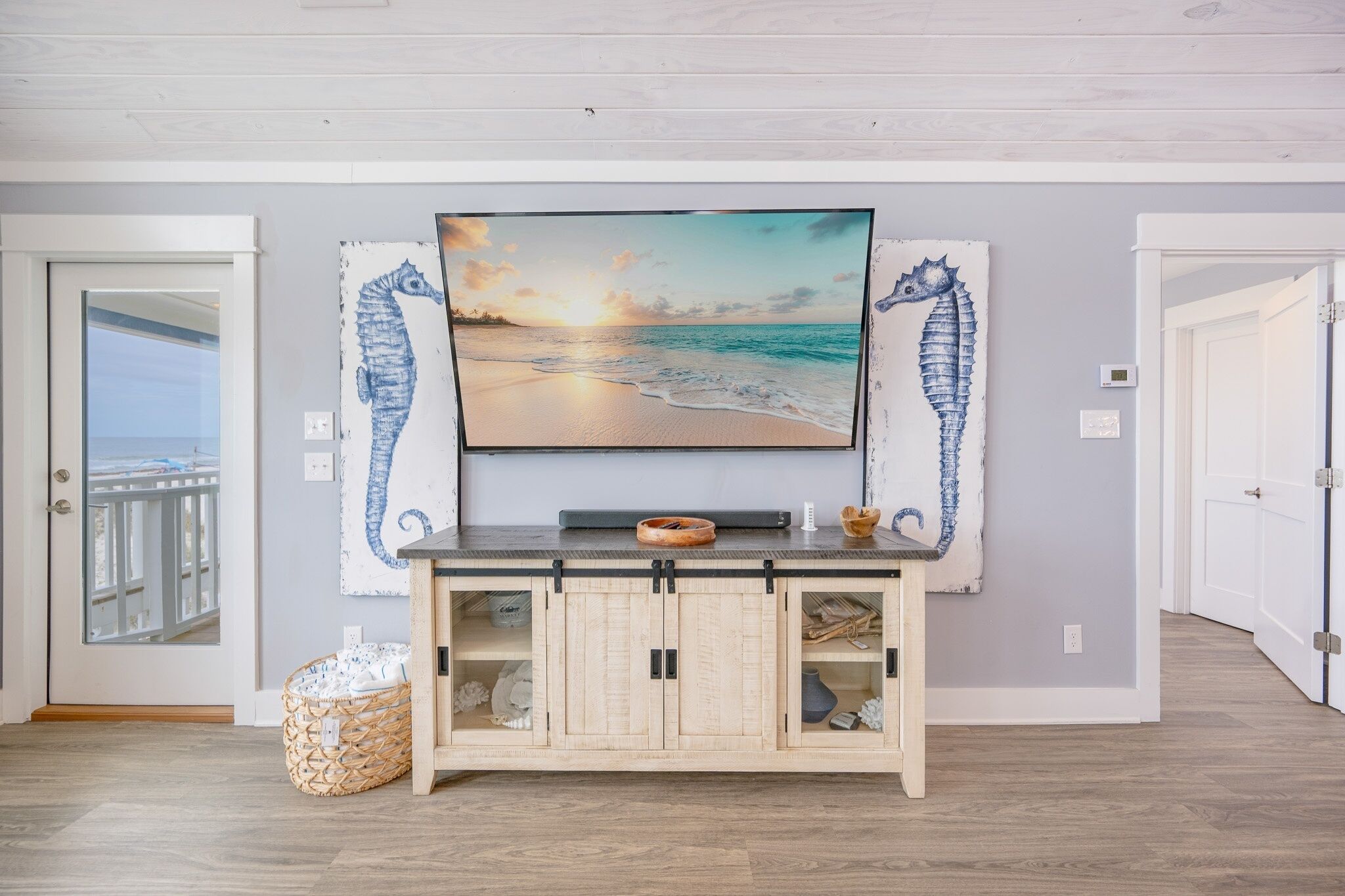 Una stanza con una grande TV montata sopra un mobile in legno, una decorazione a forma di cavalluccio marino e vista sull'oceano attraverso una porta.