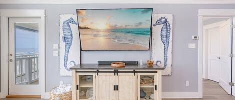 Una stanza con una grande TV montata sopra un mobile in legno, una decorazione a forma di cavalluccio marino e vista sull'oceano attraverso una porta.