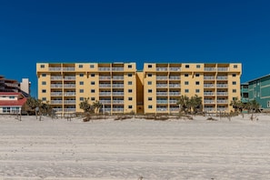 Hotel de vários andares com varandas, situado numa praia arenosa com palmeiras.