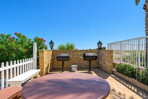 An outdoor patio with a round table and two barbecue grills, surrounded by a white fence and greenery.