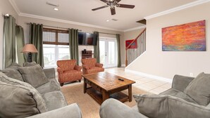 A living room with a sofa set, a coffee table, and two armchairs. There is a television mounted on the wall and a painting above the staircase. - Windswept 4 Bedroom Home (Orange Beach)
