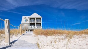 A beachfront house with a balcony, a concrete pathway leading to the entrance, and sand dunes in the background. - Windswept 4 Bedroom Home (Orange Beach)