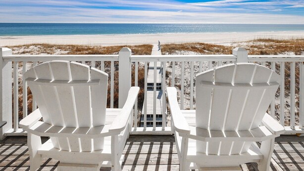 Two white Adirondack chairs facing a beach with a wooden deck and a railing. - Windswept 4 Bedroom Home (Orange Beach)