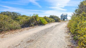A gravel road leading to a modern two-story house surrounded by dense vegetation. - Windswept 4 Bedroom Home (Orange Beach)