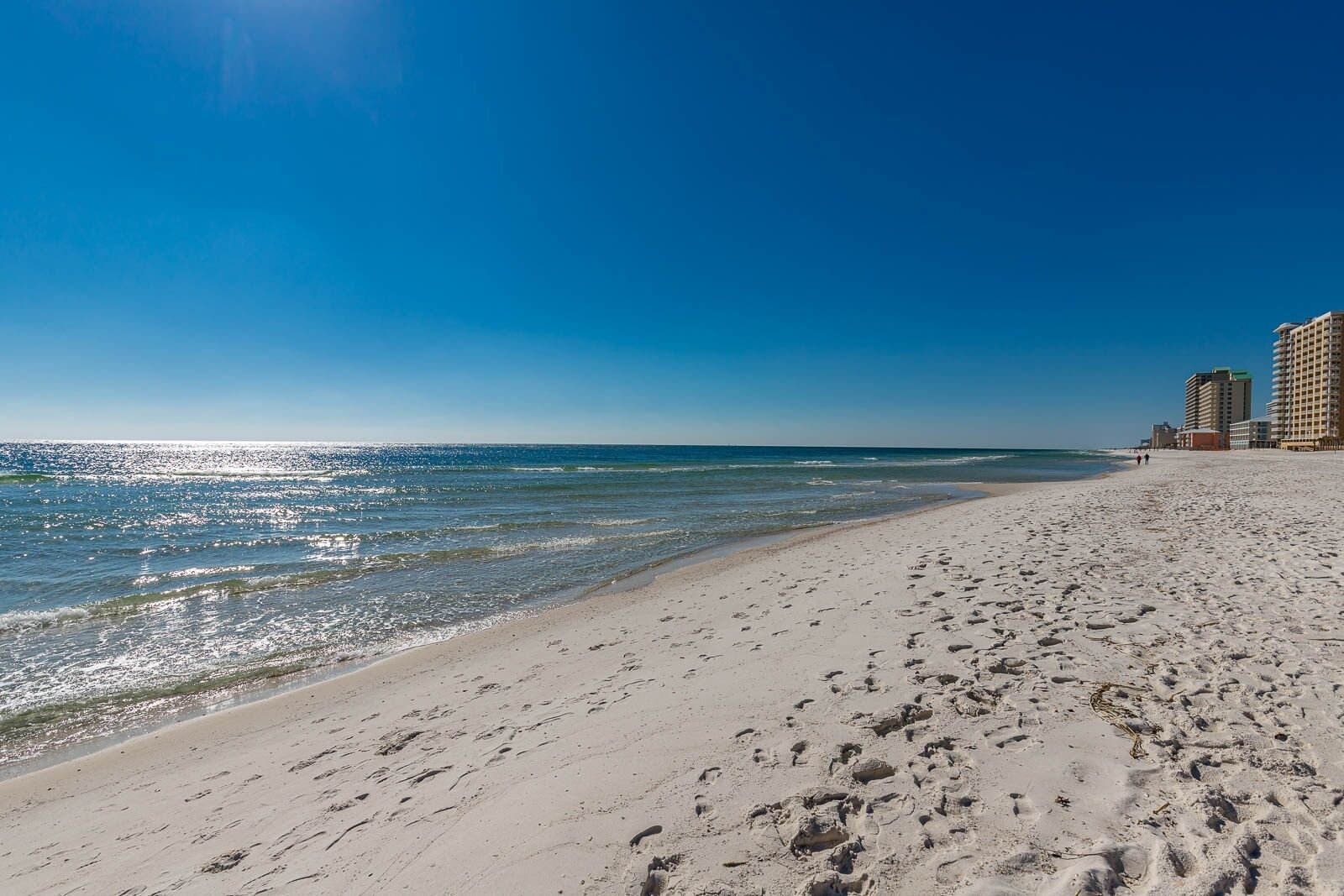 A beach with footprints, a clear sky, and buildings in the background.