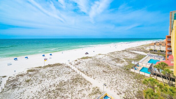 A beach with a swimming pool, beach umbrellas, and a building with a red roof. - Driftwood Towers 6d 2 Bedroom Condo (Gulf Shores)