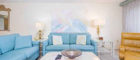 Una sala de estar con un sofá azul, una mesa de centro, un sillón amarillo y un cuadro en la pared.