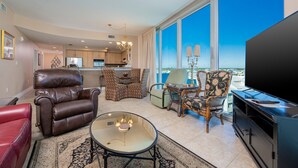A spacious living room with a leather sofa, a glass coffee table, and a large flat-screen TV. There is a dining area with a chandelier and a view of the city through large windows. - Island Tower 601 3 Bedroom Condo (Gulf Shores)