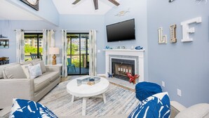 A living room with a fireplace, a ceiling fan, a flat-screen TV, and a large window with curtains. - Mariner Lakes 40 2 Bedroom Condo (Orange Beach)