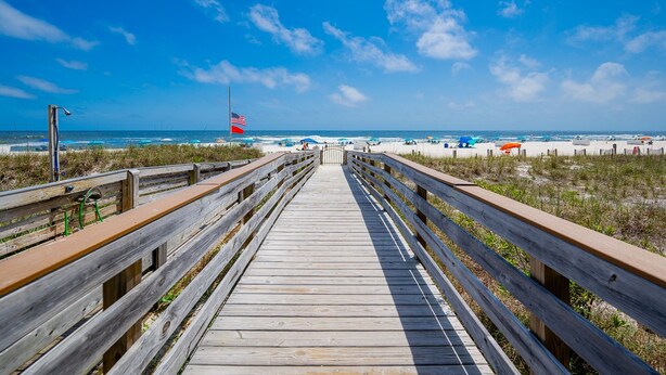A wooden boardwalk leading to a beach with a flag and umbrellas. - Mariner Lakes 40 2 Bedroom Condo (Orange Beach)