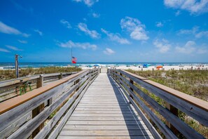 A wooden boardwalk leading to a beach with a flag and umbrellas. - Mariner Lakes 40 2 Bedroom Condo (Orange Beach)