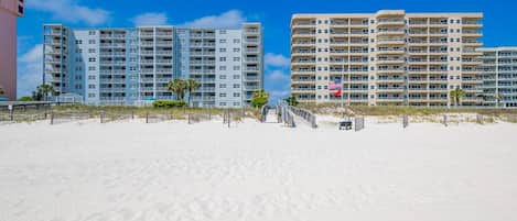 Un hotel frente a la playa con varios pisos, una playa arenosa en primer plano y un cielo azul despejado.
