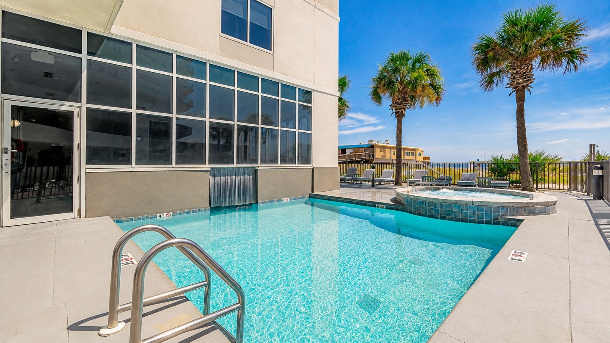 A swimming pool with a metal ladder, surrounded by a poolside area with lounge chairs and palm trees.
