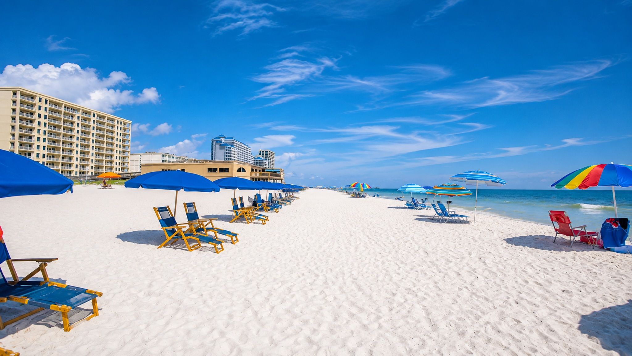 A beach with rows of sun loungers and umbrellas, a hotel building, and the ocean in the background.