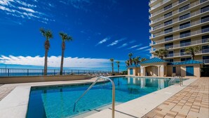 Une piscine, un ciel bleu dégagé, des palmiers et un immeuble à plusieurs étages doté de balcons.