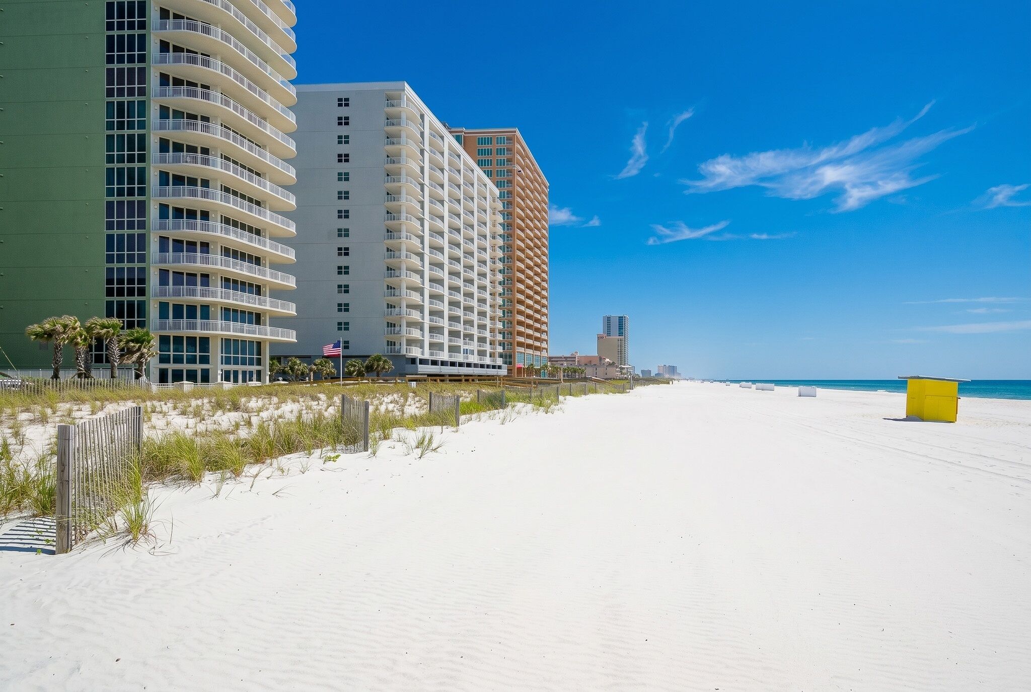 Un hotel frente a la playa con varios balcones, una playa de arena y un cielo azul despejado.