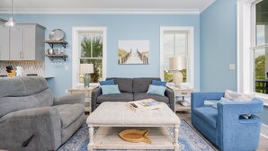 A living room with a grey sofa, a blue armchair, and a white ottoman. There is a painting on the wall and a window with a view of greenery.