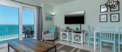 A living room with a large TV, a dining table, and chairs. There is a balcony with a view of the ocean.