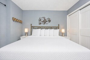 A bedroom with a bed, two bedside tables with lamps, a decorative wall piece, and a white wardrobe. - Seacrest 805 1 Bedroom Condo (Gulf Shores)