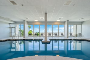 A large indoor swimming pool with clear blue water, surrounded by lounge chairs and a view of palm trees and a clear sky.
