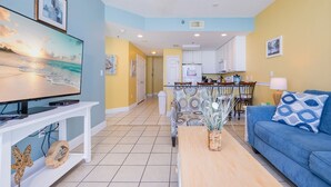 A living room with a TV, sofa, coffee table, and a view into the kitchen. - Lighthouse 1417 1 Bedroom Condo (Gulf Shores)