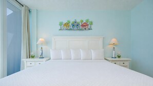 Una habitación de hotel con una cama grande, cabecera blanca, dos mesitas de noche con lámparas y un mural en la pared con casas coloridas.