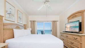 A hotel room with a large bed, a television, a ceiling fan, a balcony with curtains, and artwork on the walls. - Seawind 905 3 Bedroom Condo (Gulf Shores)