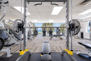 A gym with exercise equipment, a bench, and a view of palm trees through large windows. - Seawind 905 3 Bedroom Condo (Gulf Shores)