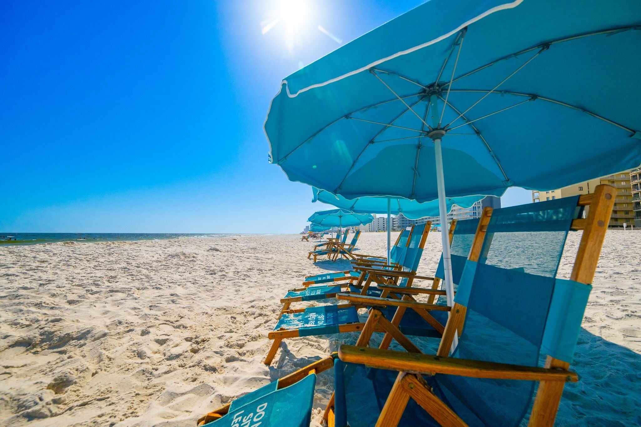 Uma praia com fileiras de cadeiras e guarda-sóis azuis, céu claro e um edifício ao fundo.
