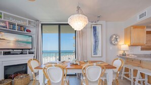 A dining area with wicker chairs, a wooden table, a chandelier, a TV, and a view of the beach. - Seaside Beach & Racquet 4407 3 Bedroom Condo (Orange Beach)
