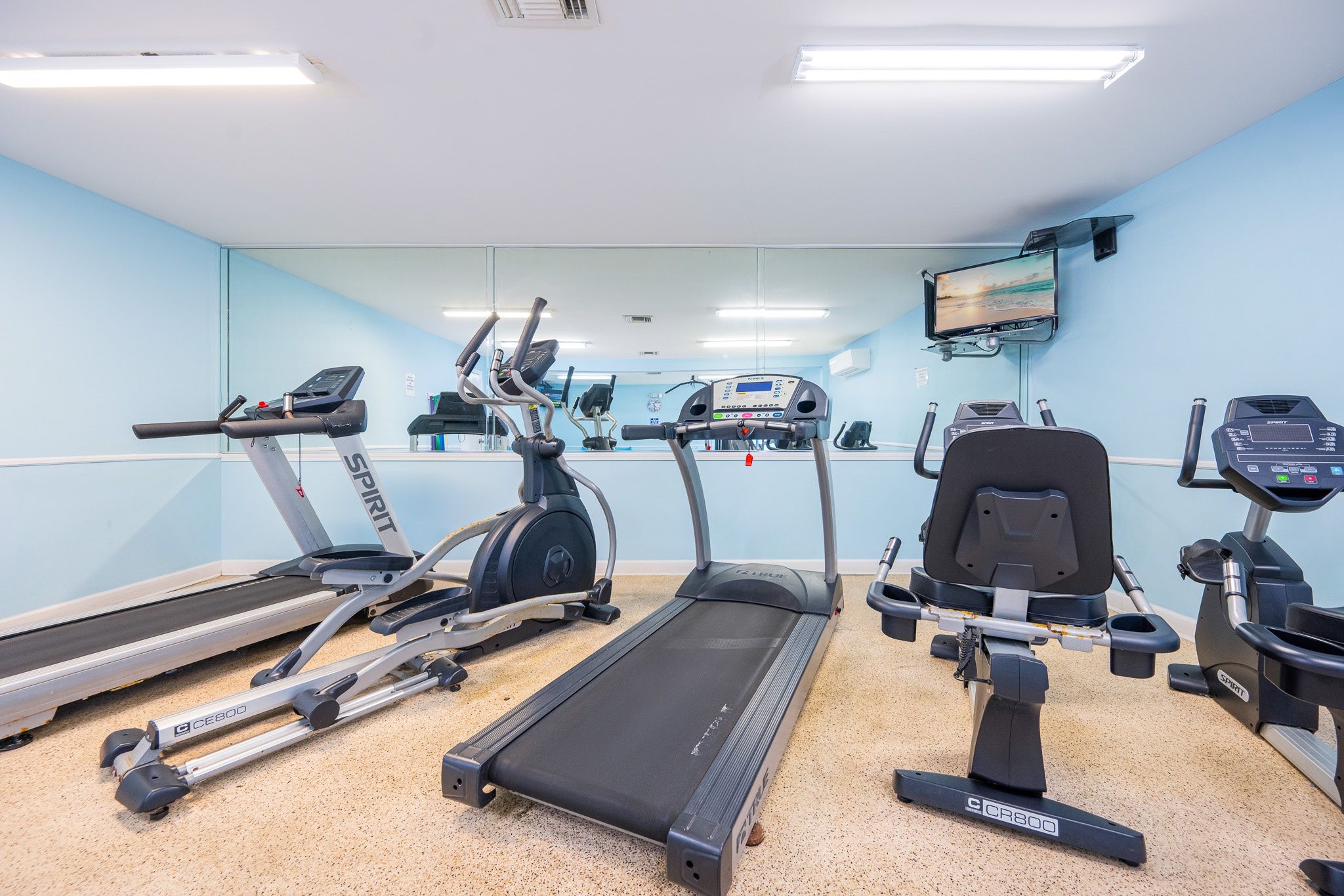 A gym with treadmills, elliptical machines, and a stationary bike, equipped with digital displays and a television on the wall.