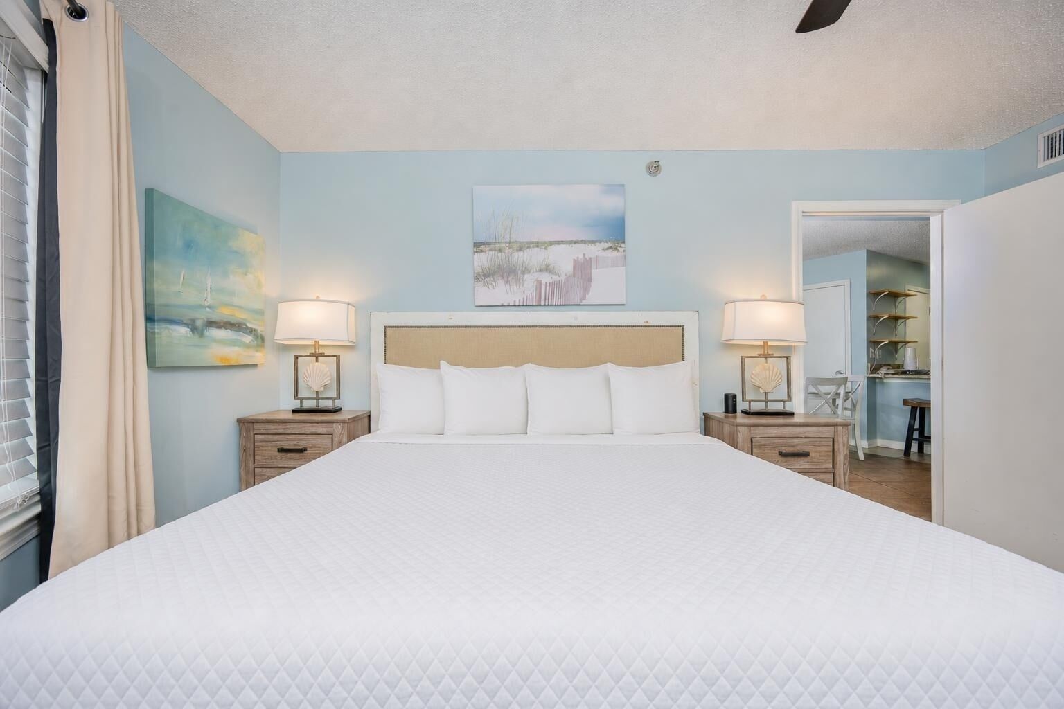 A large bed with a quilted white duvet, two bedside tables with lamps, a headboard, and a painting above the bed.