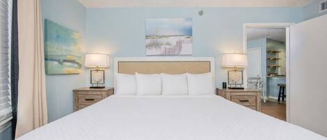 A large bed with a quilted white duvet, two bedside tables with lamps, a headboard, and a painting above the bed.
