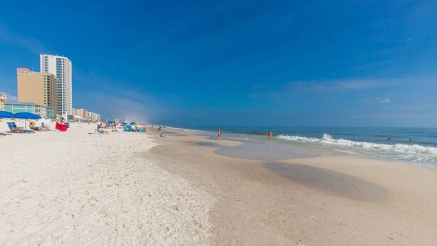 A beach with a sandy shore, a few umbrellas, and a building in the background. - Gulf Side Townhomes 20 2 Bedroom Condo (Gulf Shores)