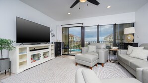 A modern living room with a flat-screen TV, a ceiling fan, and a large window with a view of buildings. - Gulf Side Townhomes 20 2 Bedroom Condo (Gulf Shores)