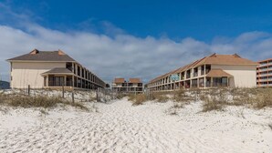 A beachfront hotel with a sandy path leading to the entrance, featuring multiple-story buildings with balconies and a clear blue sky. - Gulf Side Townhomes 20 2 Bedroom Condo (Gulf Shores)
