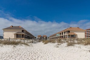 Un hôtel situé en bord de mer, avec un chemin de sable menant à l’entrée, composé d’immeubles à plusieurs étages dotés de balcons, et offrant une vue sur un ciel bleu azur.